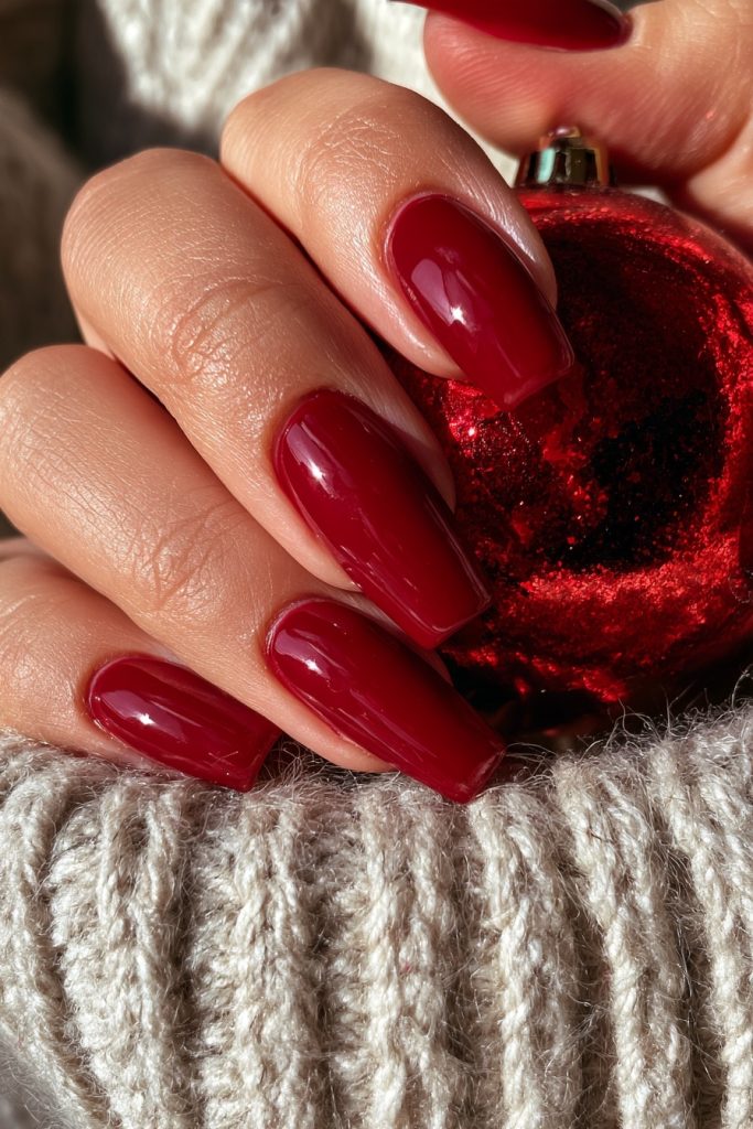 Close-up of a hand with long glossy cranberry red nails holding a red glitter Christmas ornament