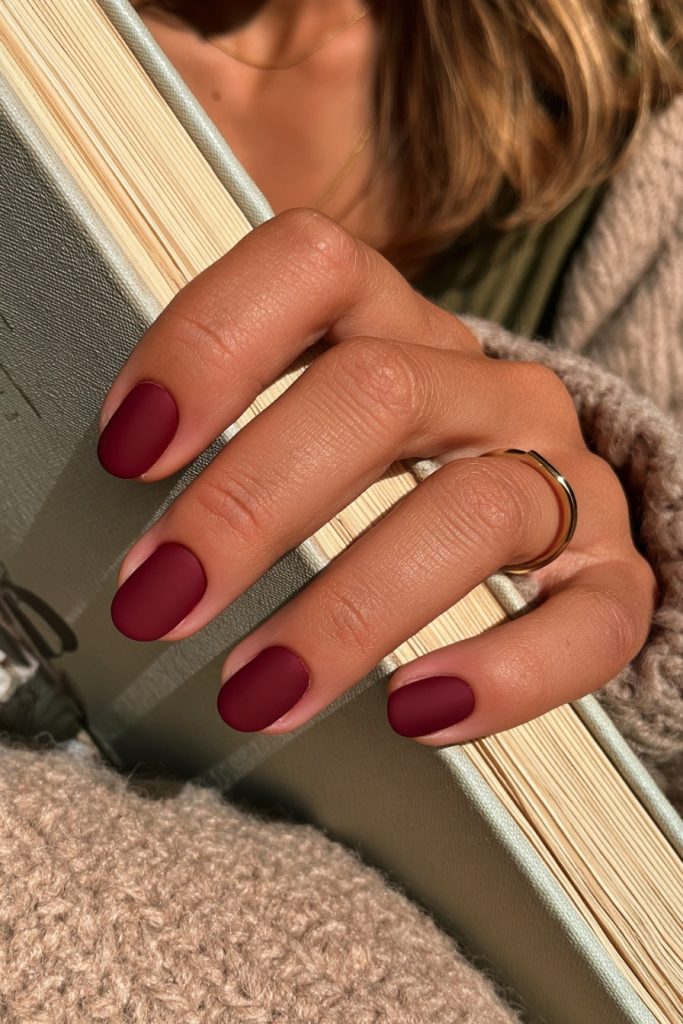 Close-up of a hand with short oval cranberry red nails and a thin gold ring resting on an open book