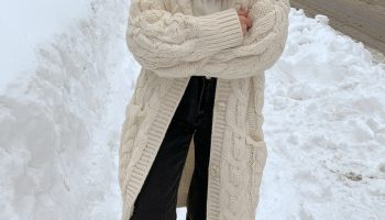 Woman in a cream cable knit cardigan over a white turtleneck with dark jeans and snow boots on a snowy street