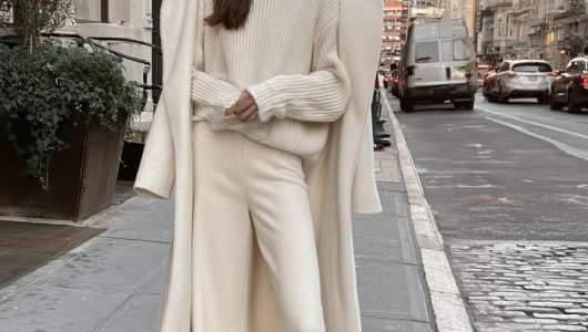Woman in a cream knit sweater and cream wide leg trousers with a long ivory wool coat worn open on a city street