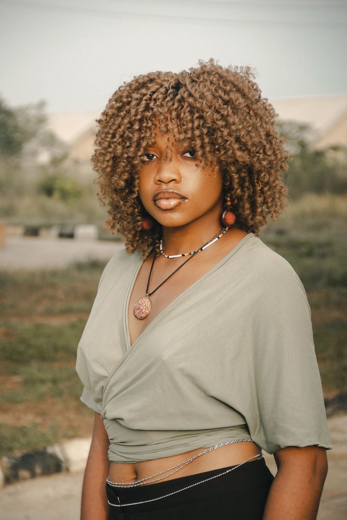 Woman with golden brown coily curls and a short curly fringe, wearing a sage green wrap top outdoors