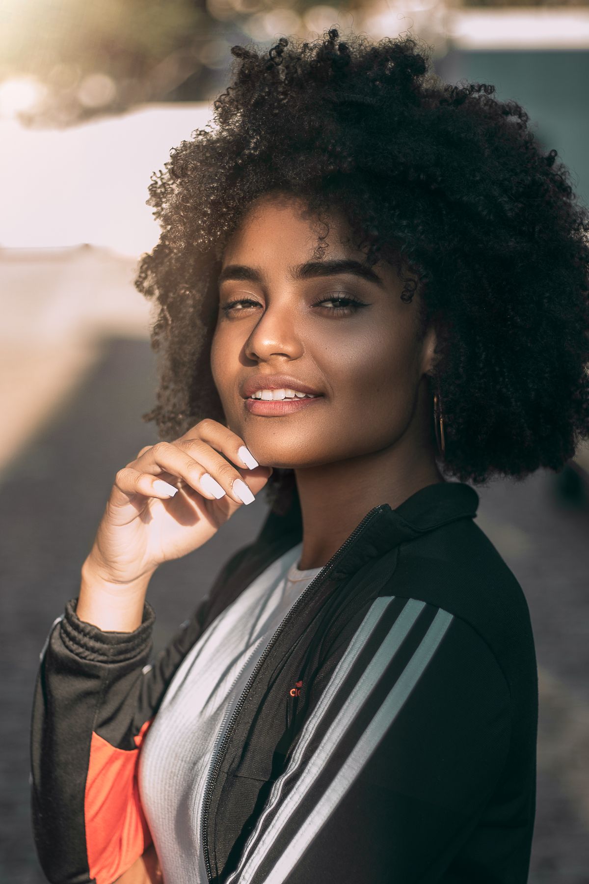 Woman with a large, rounded dark coily hairstyle with her hand at her chin, wearing a black track jacket and lit by warm golden light