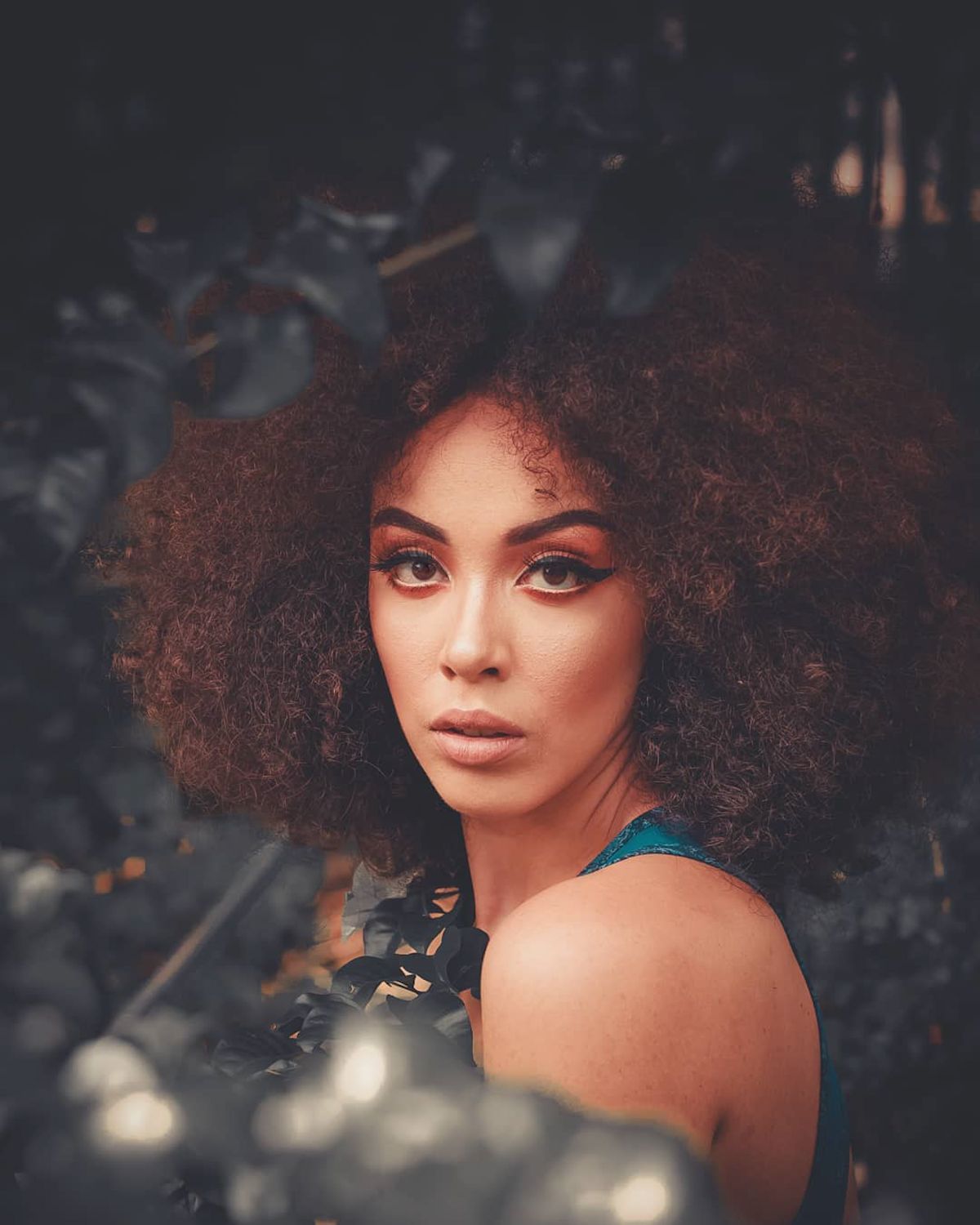 Woman with big, voluminous auburn brown curls and a teal top, photographed against dark foliage