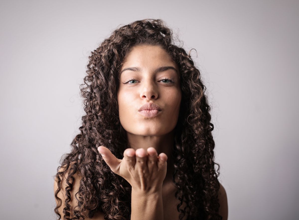 Woman with very long, dark spiral curls blowing a kiss against a light grey backdrop
