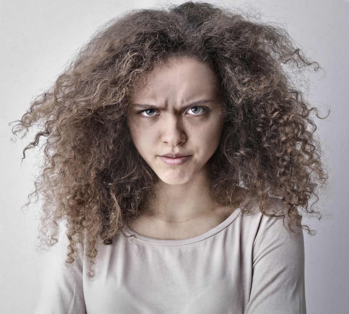 Woman with large, voluminous ash brown curly hair, a stern expression and a light grey top against a plain backdrop