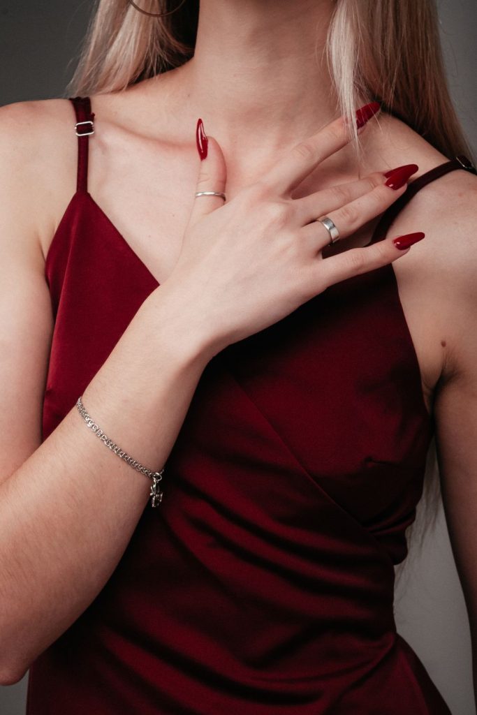 Deep red almond nails resting on a burgundy satin camisole with silver rings and a thin bracelet