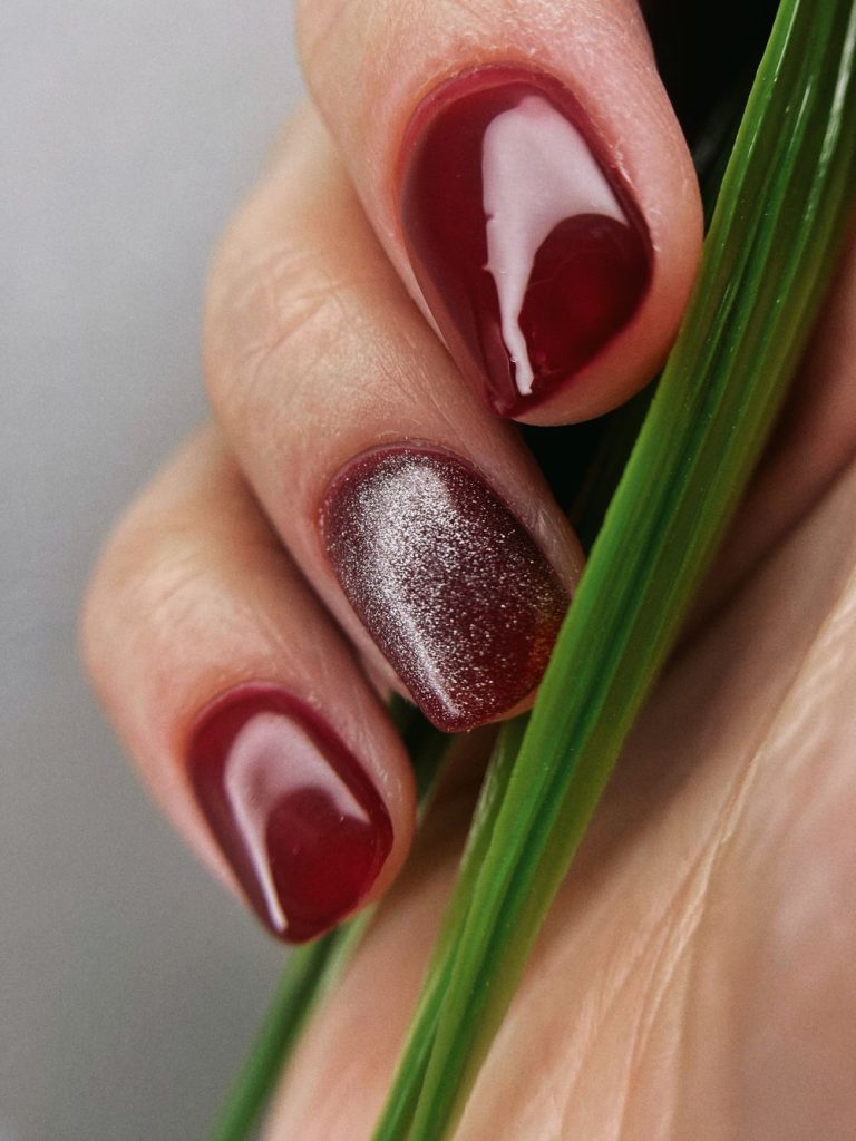 Glossy deep red gel nails with one sparkly red accent nail held beside a green blade of grass