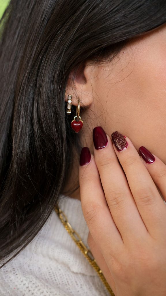 Hand with dark burgundy nails near a woman face with a cherry shaped earring and a gold chain