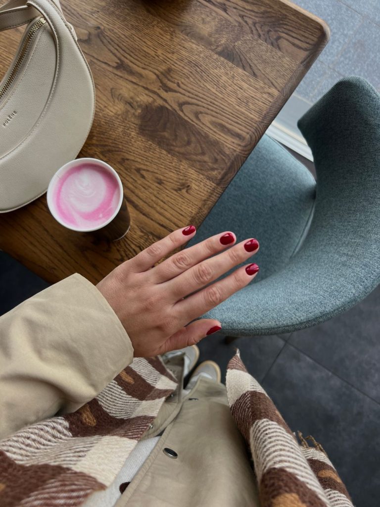 Hand with short red nails resting on a wooden cafe table next to a pink drink and a cream and brown checked sleeve