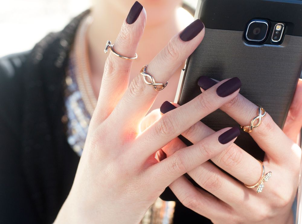 Dark plum aubergine nails with silver rings on a hand holding a phone