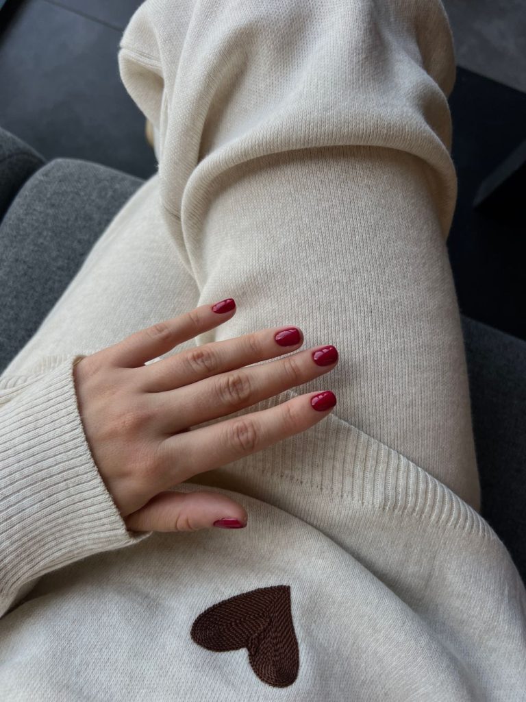 Hand with deep red oval nails resting on a cream knit sweater that has a small brown heart patch