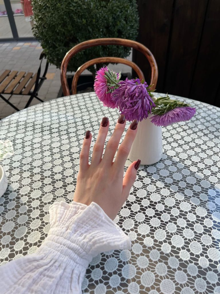 Hand with red nails reaching toward purple flowers on a lace patterned cafe table, with a white ruffled cuff