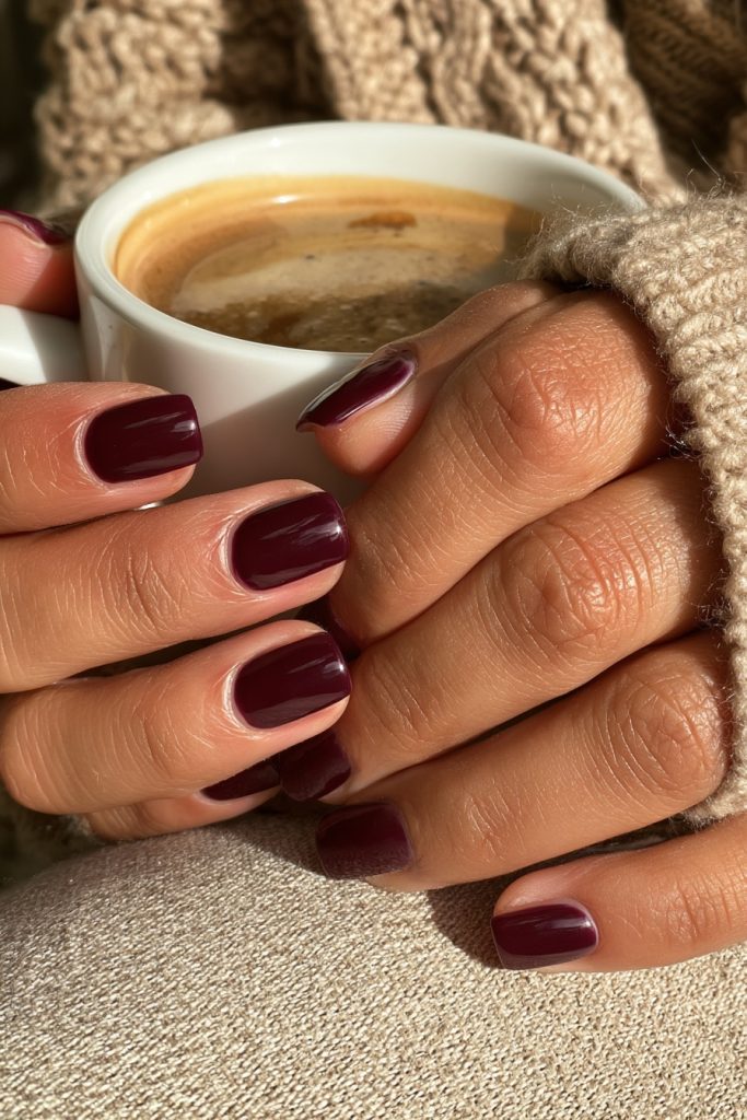 Close up of hands with deep plum nails holding a white coffee cup in a cozy knit sweater
