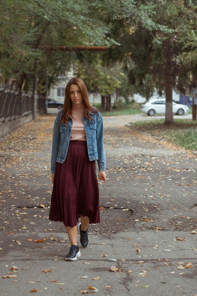 Woman in a mid-wash denim jacket, a pale top, a long burgundy skirt and dark sneakers walking along an autumn path