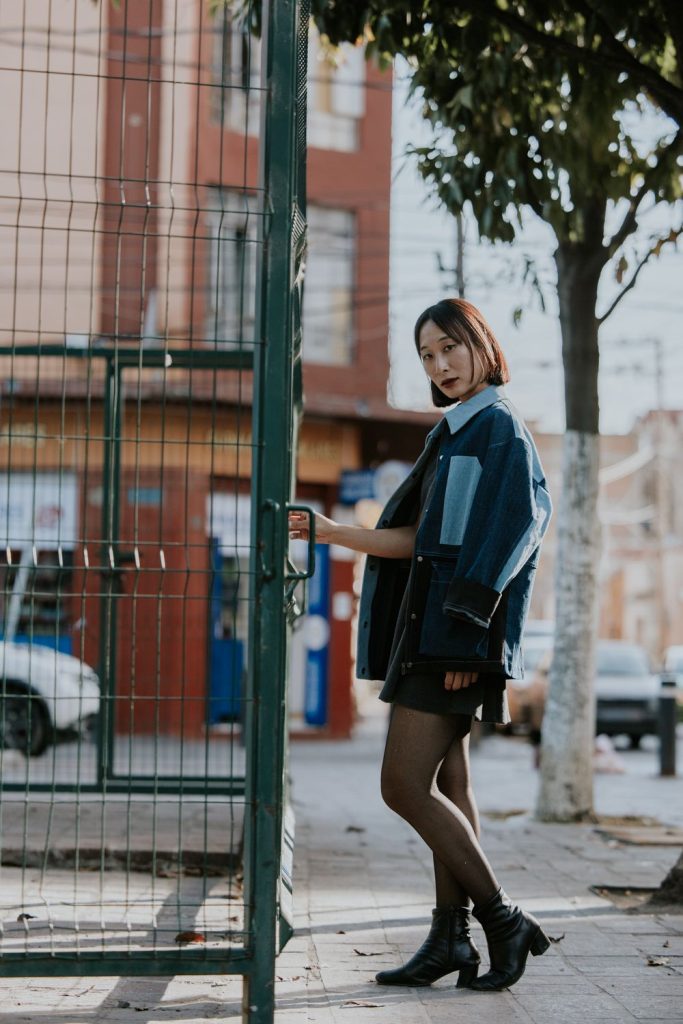 Woman by a green gate in a two-tone patchwork denim jacket in dark and light blue with a black skirt, tights and black boots