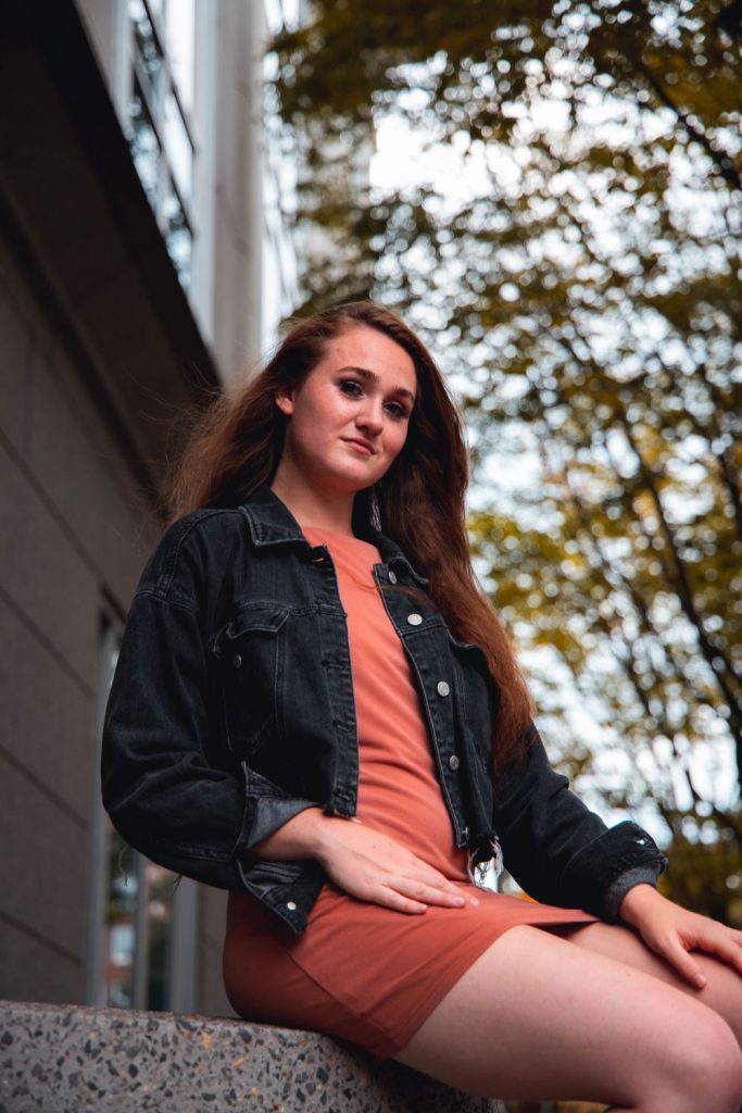 Woman with long red-brown hair in a black denim jacket over a rust orange dress sitting outdoors