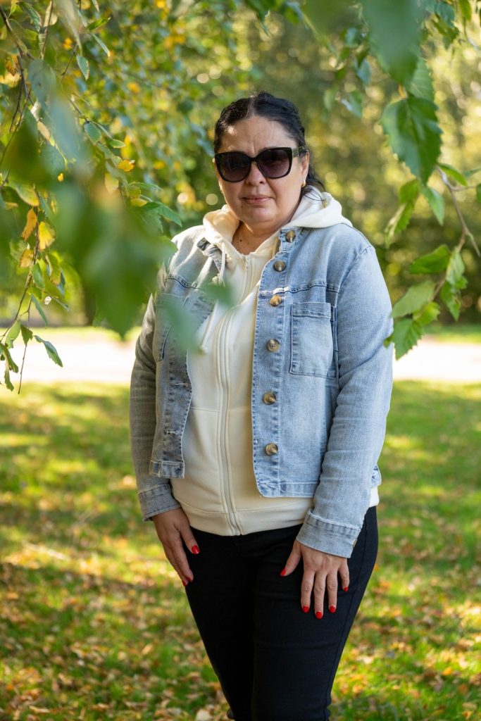Woman in sunglasses wearing a light blue denim jacket over a cream hoodie and black trousers in a sunny green field