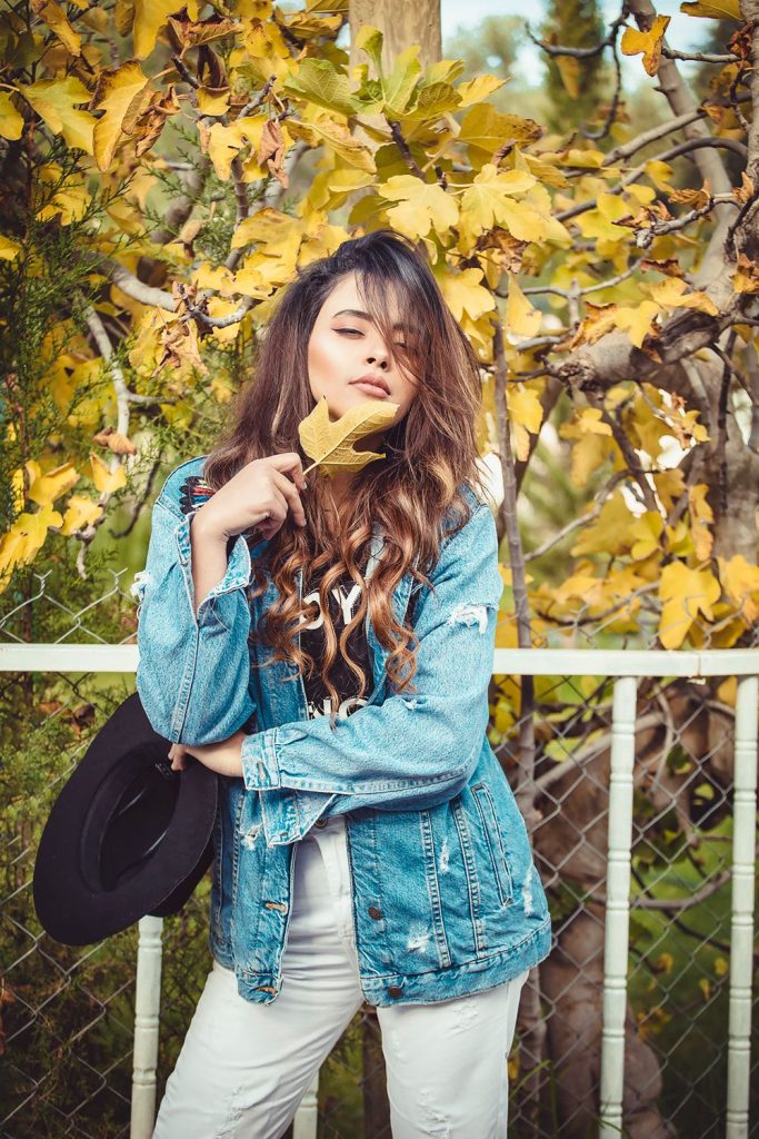 Woman with long wavy hair in a distressed light blue denim jacket and white trousers holding a black wide-brim hat and a yellow leaf
