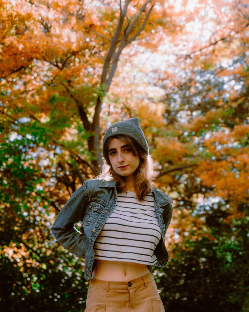 Woman in a grey beanie, a washed denim jacket, a striped top and mustard trousers among orange autumn trees