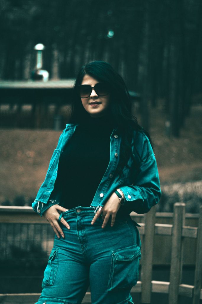 Woman in sunglasses and a black hat wearing a teal denim jacket, a black turtleneck and teal jeans leaning on a fence