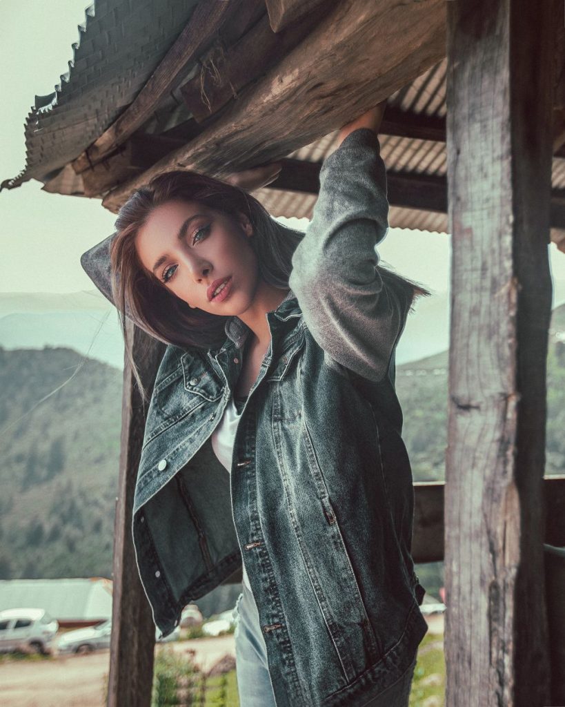 Woman with long brown hair leaning against a wooden post in an oversized distressed grey-blue denim jacket