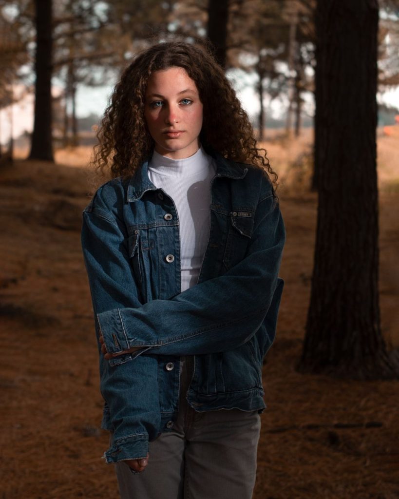Woman with long curly hair in a dark denim jacket over a white turtleneck standing in a forest