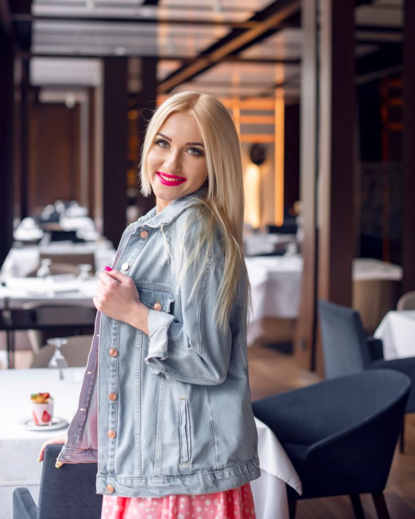 Smiling woman with long blond hair in a light denim jacket and a pink skirt standing in a restaurant