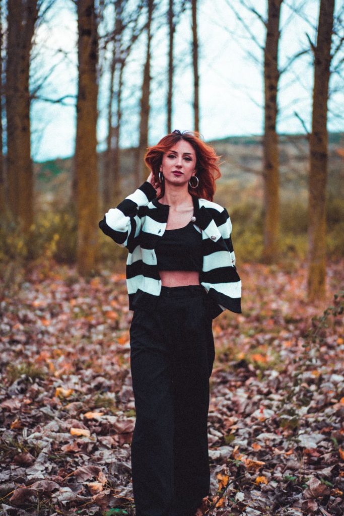 Woman with red hair in a black and pale striped cropped cardigan, a black crop top and a long black skirt among autumn leaves