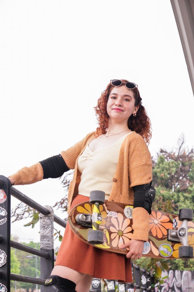 Young woman in an orange cardigan, a cream top and a rust skirt holding a floral skateboard