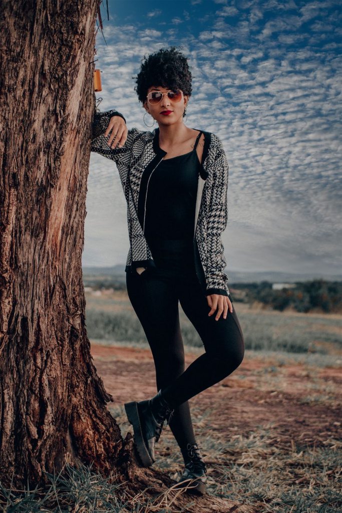 Woman with short curly hair and sunglasses in a black and white checked cardigan, a black top, black trousers and boots leaning on a tree