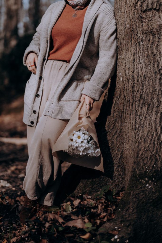 Woman leaning on a tree in a grey cardigan, a rust orange top and a long cream skirt carrying a small floral bag