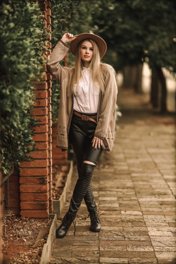 Woman in a brown hat, a long taupe cardigan, a white blouse, black leather trousers and tall black boots on a brick path