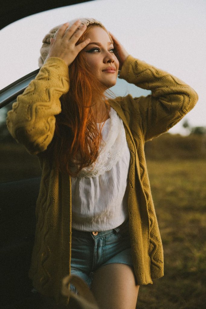 Woman with red hair in a mustard cable knit cardigan, a white lace camisole and denim shorts in golden light