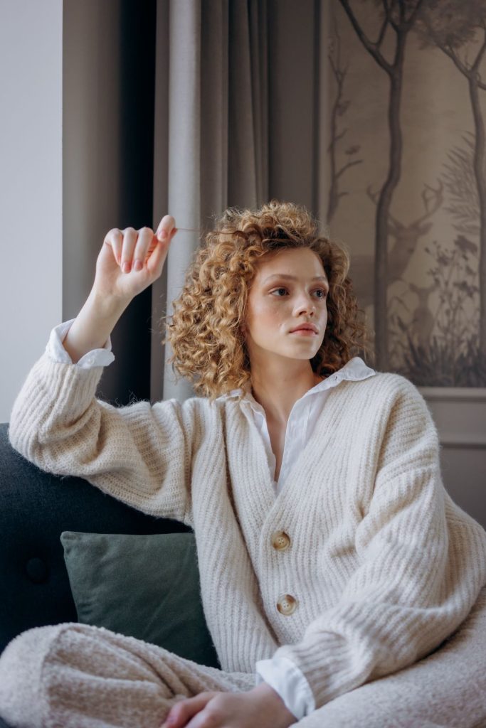Woman with curly hair in a cream chunky collared cardigan with buttons sitting on a sofa