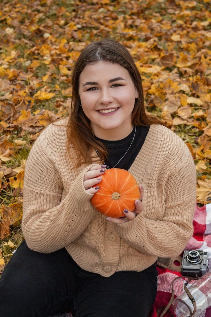 Smiling woman in a camel cardigan over a black top holding a small orange pumpkin among autumn leaves