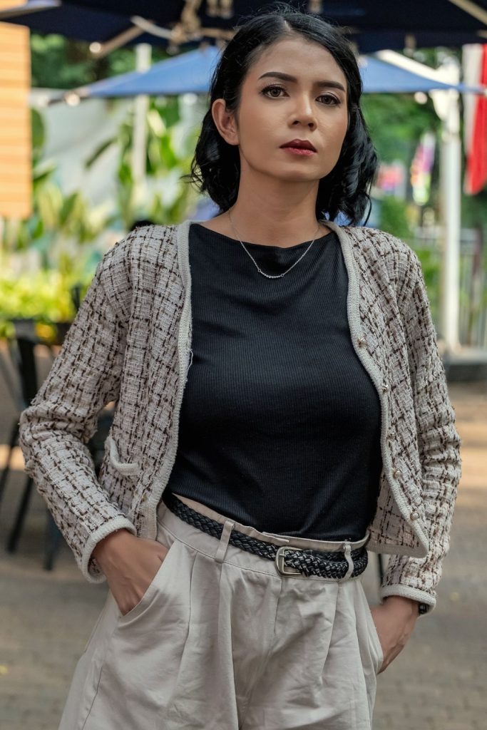 Woman in a grey and black tweed cardigan, a black tee, a thin gold necklace and cream pleated trousers with a woven belt
