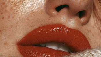 Close up of lips with a glossy burnt orange lipstick and golden freckles beside a knit sleeve