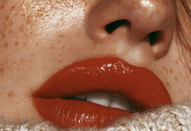 Close up of lips with a glossy burnt orange lipstick and golden freckles beside a knit sleeve