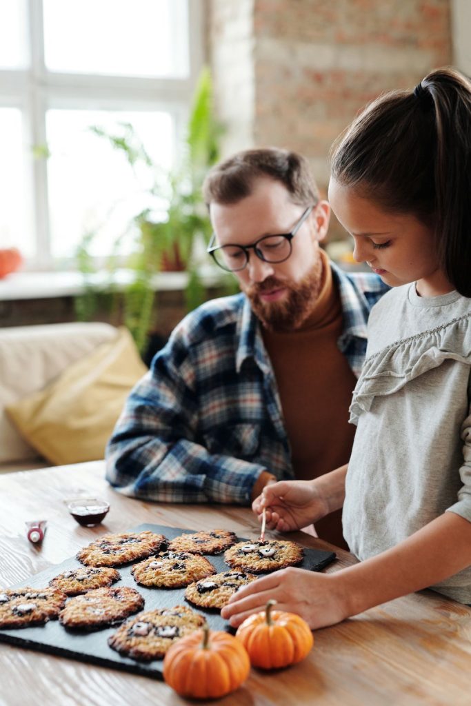 Father and children decorating round cookies at a kitchen table beside small orange pumpkins