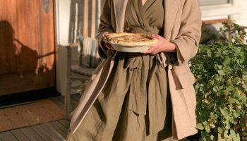 Woman in an olive green wrap dress with a camel blazer and brown boots holding a pie on a porch