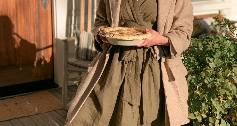 Woman in an olive green wrap dress with a camel blazer and brown boots holding a pie on a porch