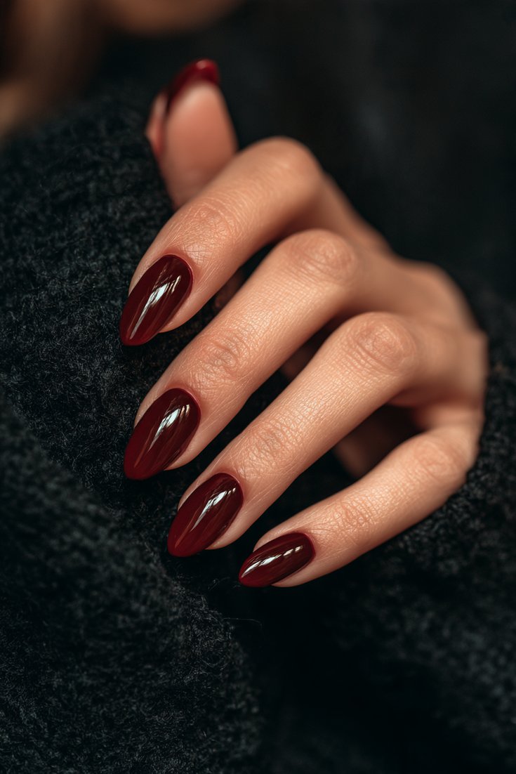 Left hand with short almond Gel X nails in deep glossy cherry cola red resting on a black wool sweater