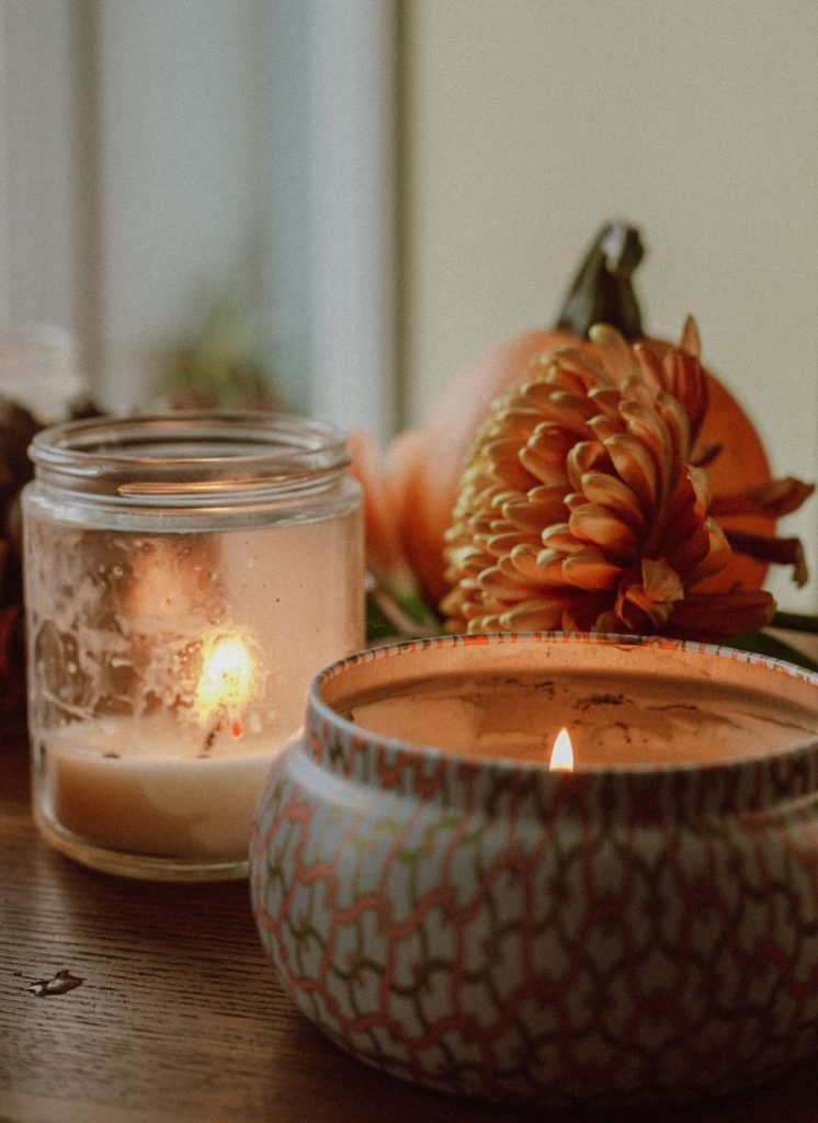 Glass jar candle glowing beside a tin candle, an orange chrysanthemum and a small pumpkin on a wooden table
