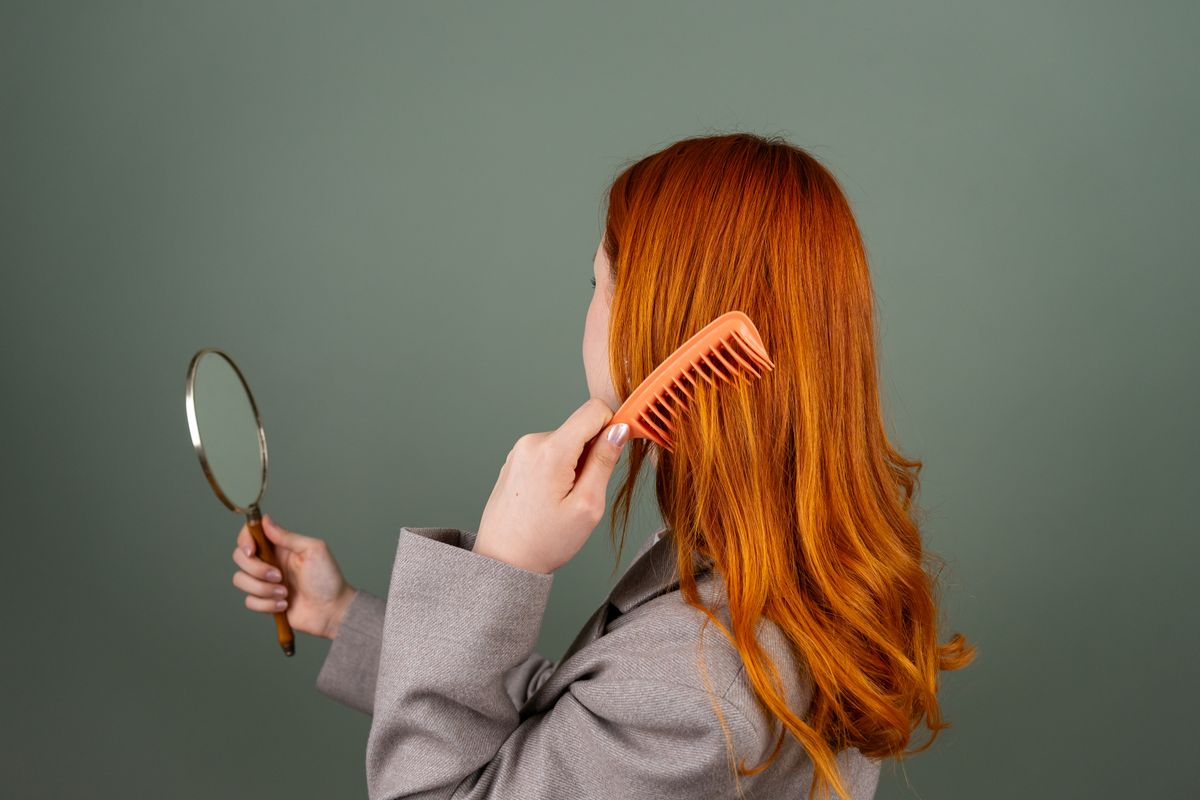 Woman with long copper red hair seen from behind, holding a comb and a hand mirror, wearing a grey jacket in front of a sage green wall