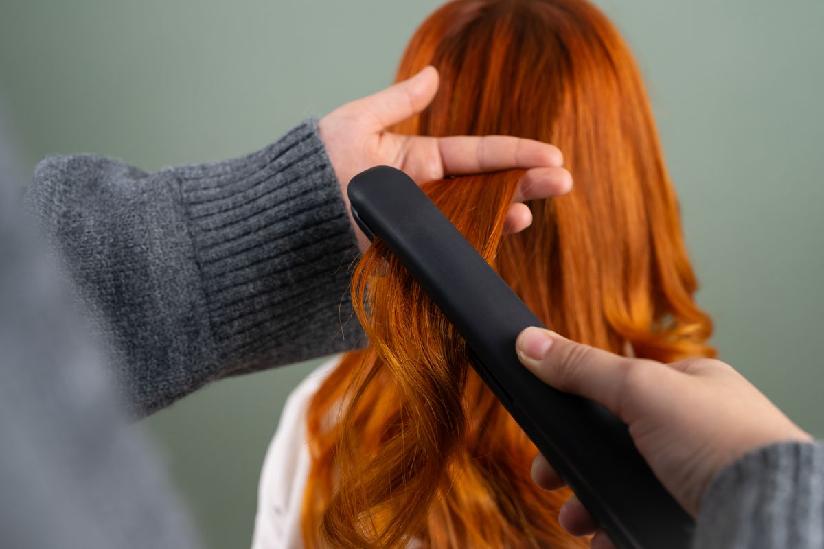 Hands in a grey sweater pressing a flat iron down a section of long copper red hair