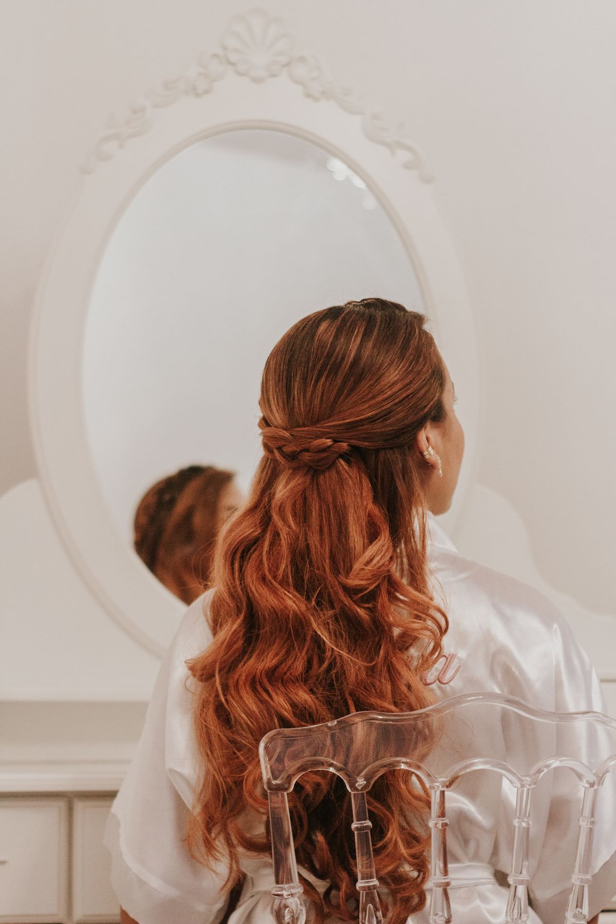 Woman seen from behind sitting in front of an oval mirror in a white satin robe, her long copper red waves half pinned up with a small braid across the back