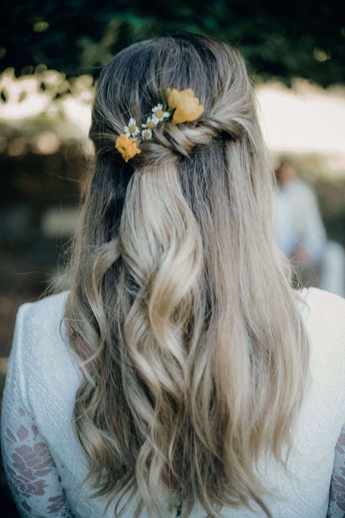 Woman seen from behind with long ash blonde waves, a half up twist decorated with small yellow and white flowers, wearing a pale blue lace jacket outdoors