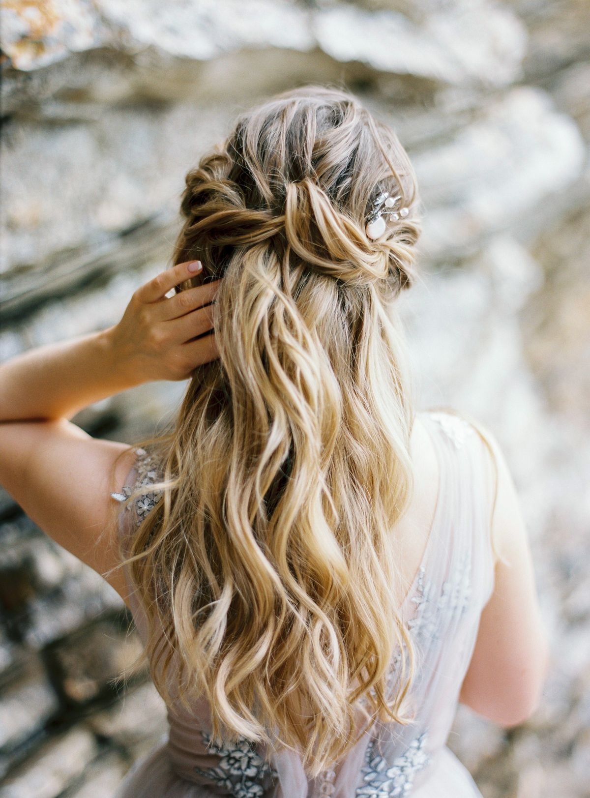 Woman seen from behind with long wavy blonde hair half up in a loose twist, one hand touching her hair, wearing a grey lilac beaded gown against a rocky wall