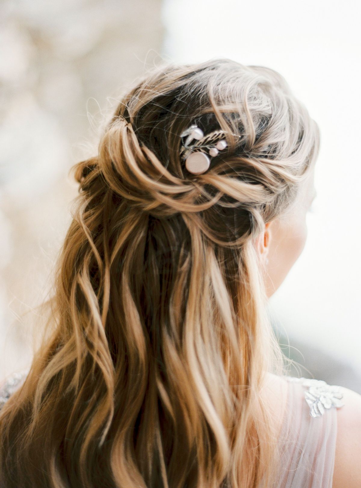 Woman seen from behind with honey blonde wavy hair twisted half up and held with a small silver hair pin, wearing a light tulle dress with beading