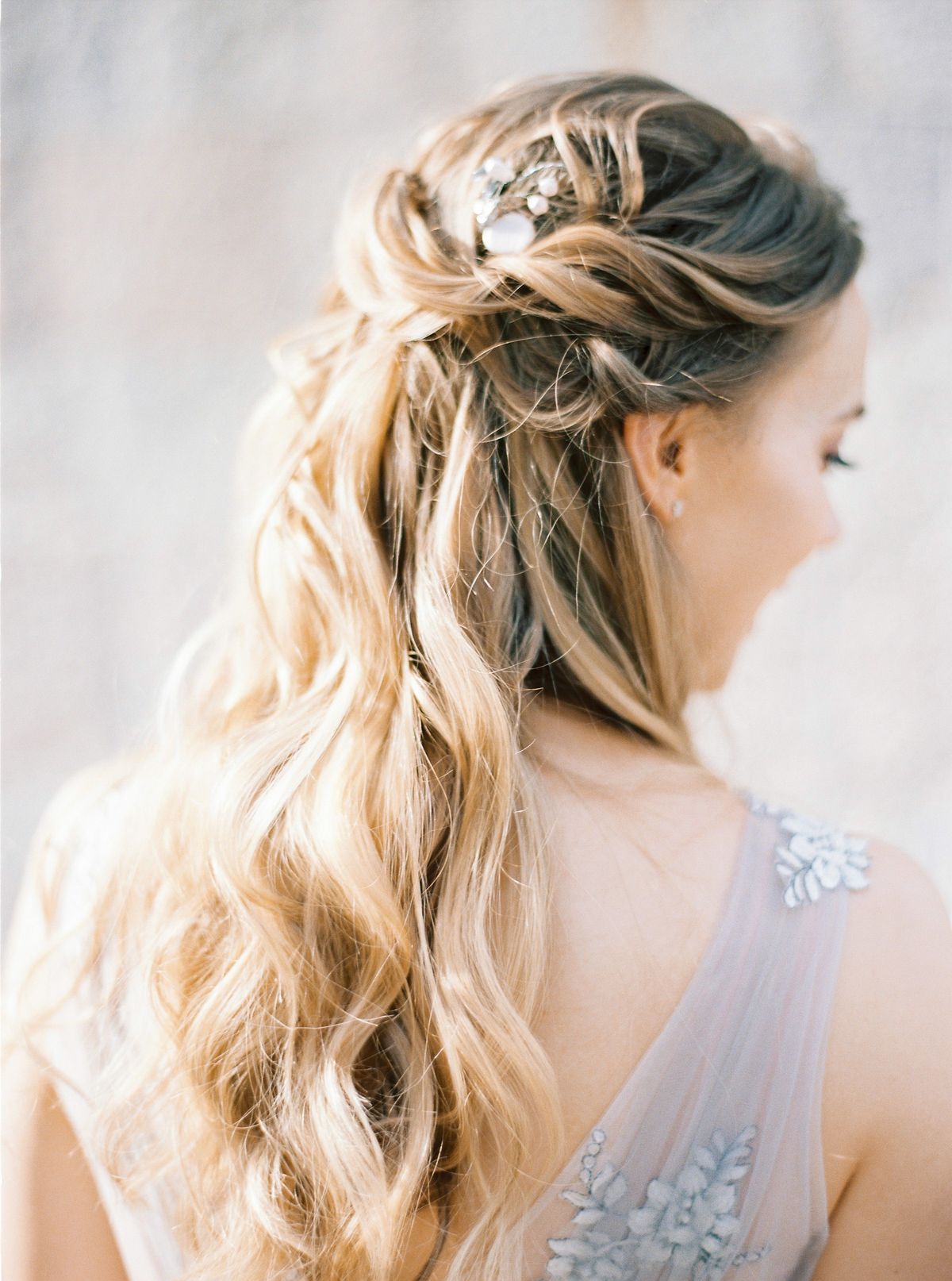 Woman in three quarter view with long wavy honey blonde hair, a twisted half up section and a small silver hair pin, wearing a light dress with beaded straps